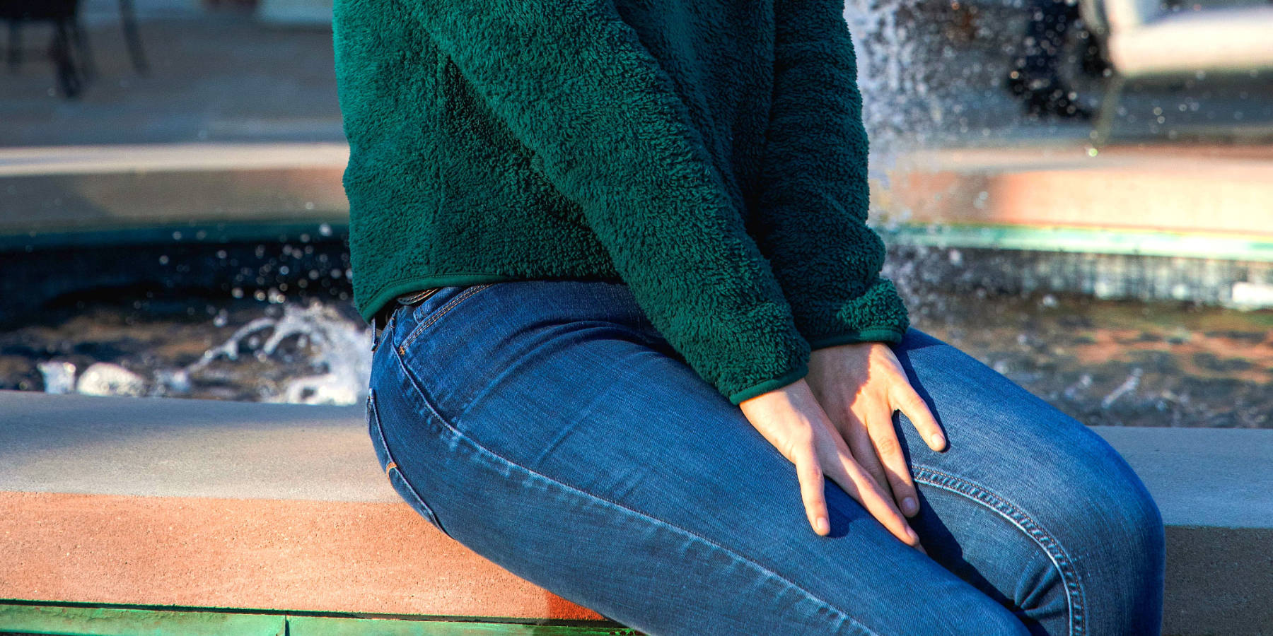 Junge Frau in Jeans sitzt am Brunnen und pinkelt sich öffentlich in die Hose.