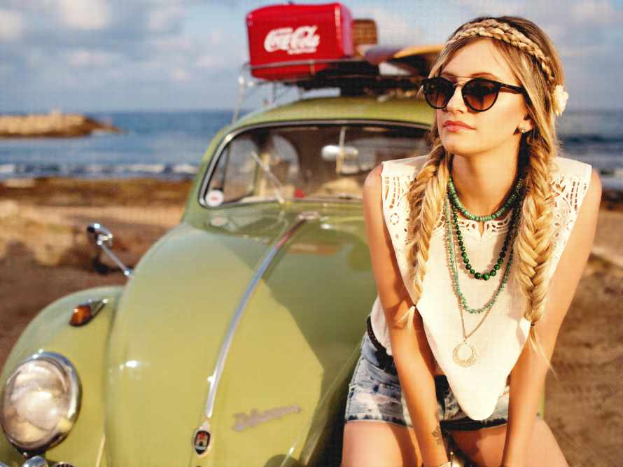 Junge Frau im Hippie-Look lehnt am Strand an einem VW Käfer