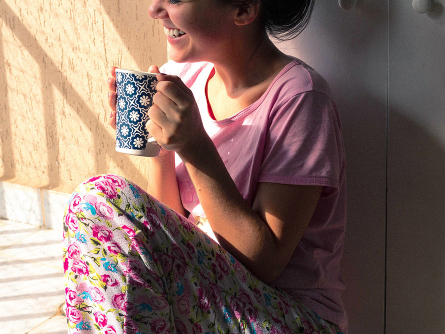 Junge Frau im Pyjama sitzt mit einer Tasse Kaffee auf dem Balkon in der Morgensonne