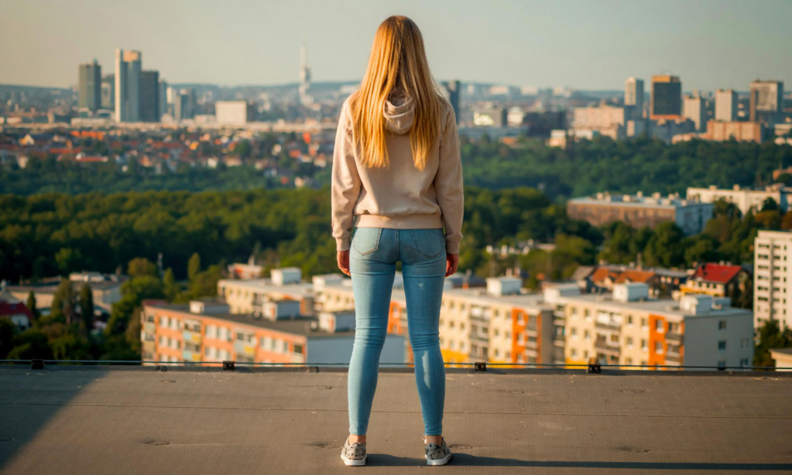 Symbolbild (Pexels, modifiziert mit KI-Unterstützung): Junge, blonde Frau steht auf dem Flachdach eines Hochhauses und macht sich, während sie in die Ferne schaut, mit voller absichtlich ihr großes Geschäft in die Hose.