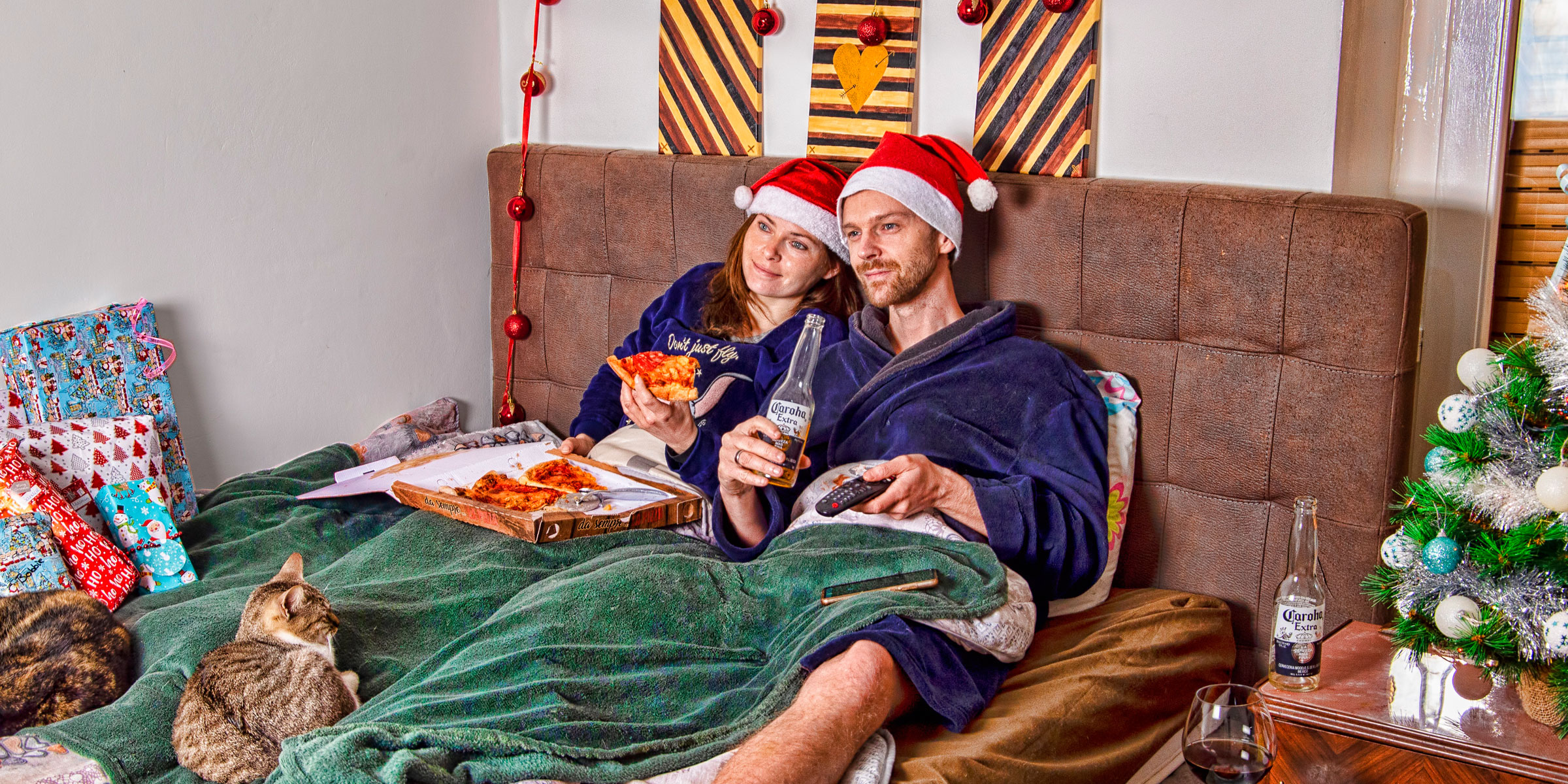 Junges Paar liegt umgeben von Geschenken mit Weihnachtsmützen im Bett und isst Pizza